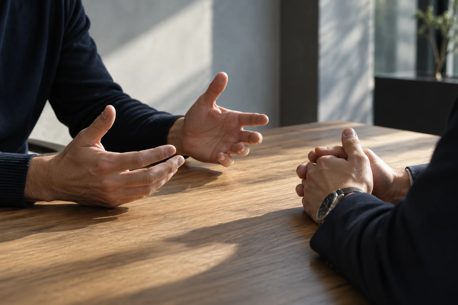 Two people's hands across a wooden table, one pair gesturing to explain and the other clasped, listening